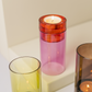 Ein rosa-roter Kerzenhalter aus Glas, das „Remember Windlicht ‚Lys‘ Pink“ von Remember, besticht durch ein auffälliges Colour-Blocking-Design und steht mit einer brennenden Kerze darin neben gelb- und brauntönigen Glasvasen auf einer hellen Unterlage. Dieses Windlicht ist mundgeblasen und handgefertigt und verleiht Ihrer Einrichtung handwerklichen Charme.
