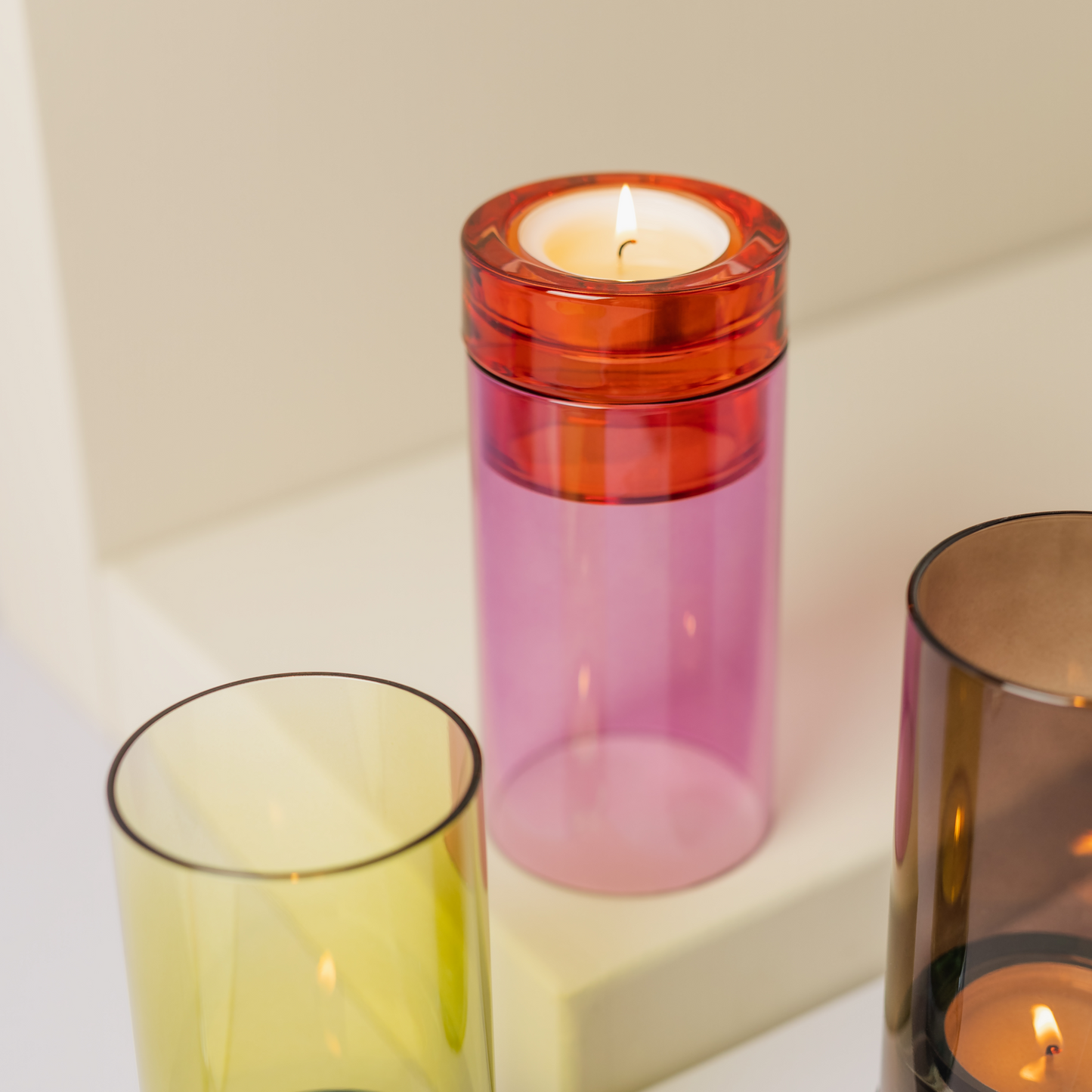 Ein rosa-roter Kerzenhalter aus Glas, das „Remember Windlicht ‚Lys‘ Pink“ von Remember, besticht durch ein auffälliges Colour-Blocking-Design und steht mit einer brennenden Kerze darin neben gelb- und brauntönigen Glasvasen auf einer hellen Unterlage. Dieses Windlicht ist mundgeblasen und handgefertigt und verleiht Ihrer Einrichtung handwerklichen Charme.