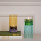 Zwei Kerzenhalter aus Glas, darunter das „Remember | Windlicht ‚Lys‘“ in Flieder von Remember, einer in Gelb- und Violetttönen und der andere in Grün- und Blautönen, stehen auf einer weißen Fläche. Der gelbe Kerzenhalter ruht auf einem Stapel Bücher mit den Aufschriften „ROM“ und „BENS“; beide Kerzen brennen und werden von einer weißen Wand im Hintergrund eingerahmt.