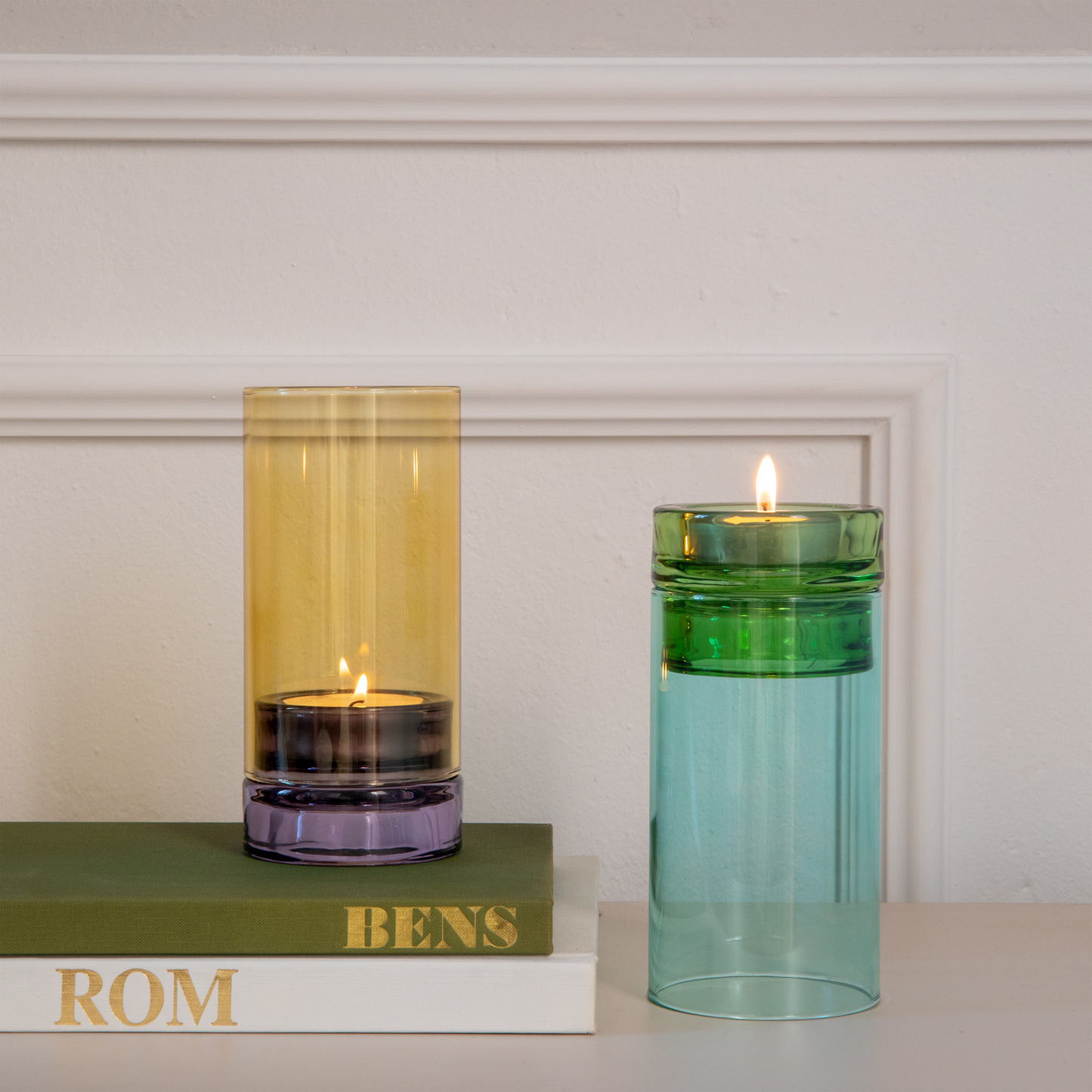 Zwei Kerzenhalter aus Glas, darunter das „Remember | Windlicht ‚Lys‘“ in Flieder von Remember, einer in Gelb- und Violetttönen und der andere in Grün- und Blautönen, stehen auf einer weißen Fläche. Der gelbe Kerzenhalter ruht auf einem Stapel Bücher mit den Aufschriften „ROM“ und „BENS“; beide Kerzen brennen und werden von einer weißen Wand im Hintergrund eingerahmt.