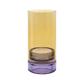 Das „Remember | Windlicht ‚Lys‘“ in Flieder von Remember ist ein zylindrischer Kerzenhalter aus Glas mit einem gelben Oberteil und einem violetten Sockel, in dem eine kleine Teelichtkerze steckt.