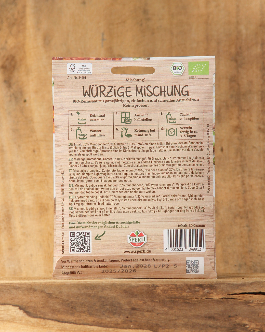 Eine braune Sperli's-Packung mit der Aufschrift "Bio Keimsprossen Saatgut 'Würzige Mischung'" steht vor einem hölzernen Hintergrund. Auf der Vorderseite stehen Produktinformationen und Anleitungen in deutscher Sprache, unten befindet sich ein Strichcode - ideal für Ihre Sprossenbox.