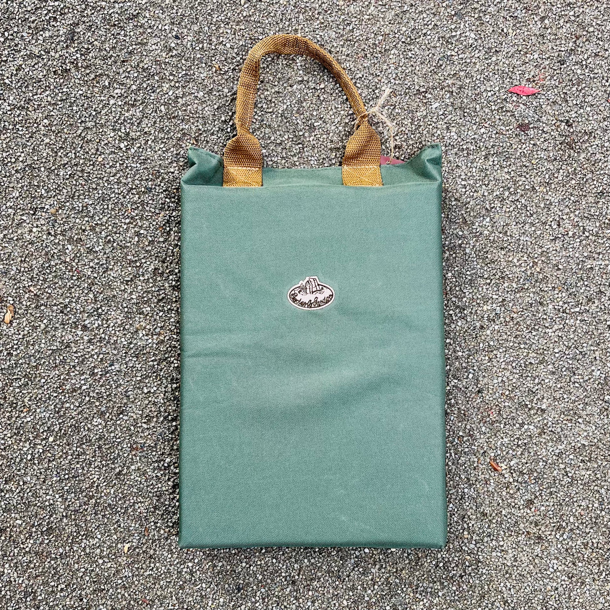 Das Esschert | Kniekissen von Eschert Design ist eine grüne, rechteckige Tasche mit braunen, gewebten Griffen, ideal für die Gartenarbeit. Es hat einen kleinen ovalen Aufnäher mit Text in der Mitte und ruht auf einer strukturierten grauen Oberfläche.