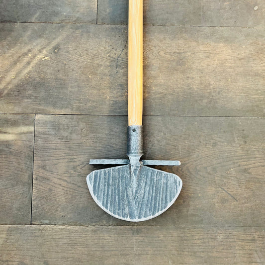 Ein Krumpholz Rasenkantenstecher 85cm mit einem Holzgriff und einer halbrunden, geriffelten Metallklinge mit zwei kurzen horizontalen Stäben liegt flach auf einem Holzboden.