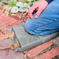 Eine Person in Blue Jeans kniet auf einem Esschert | Kniekissen von Eschert Design, einem grauen Gartenkniekissen mit braunen Griffen, das bei der Gartenarbeit auf einem Ziegelweg neben grünen Blattpflanzen platziert wird.